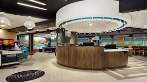 A modern hotel lobby with a central reception desk, seating areas, and a large mural on the wall.