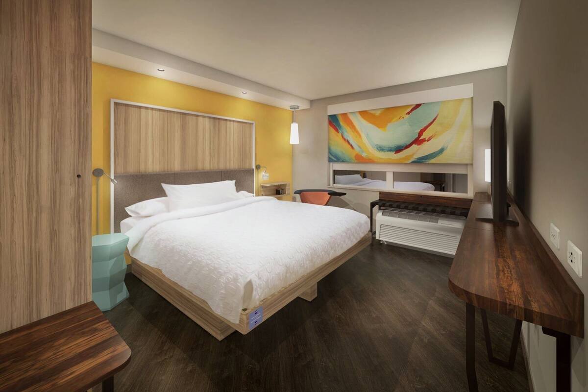 A modern hotel room with a large bed, a desk, a chair, a TV, and a colorful abstract painting on the wall.