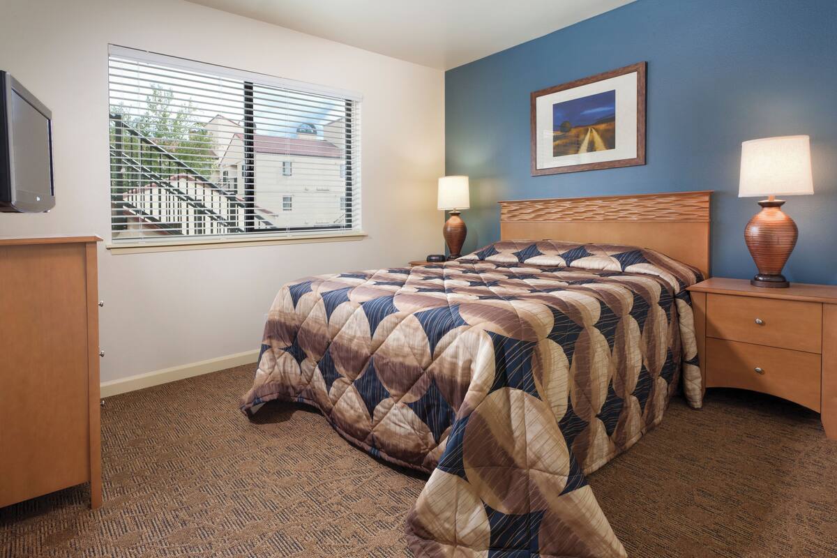 A bedroom with a bed, two bedside tables with lamps, a dresser, a window with blinds, and a framed picture on the wall.