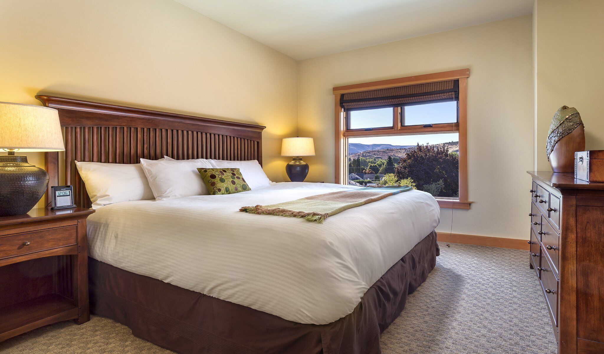 A neatly made bed with a wooden headboard, two bedside lamps, a digital clock, and a dresser with drawers.