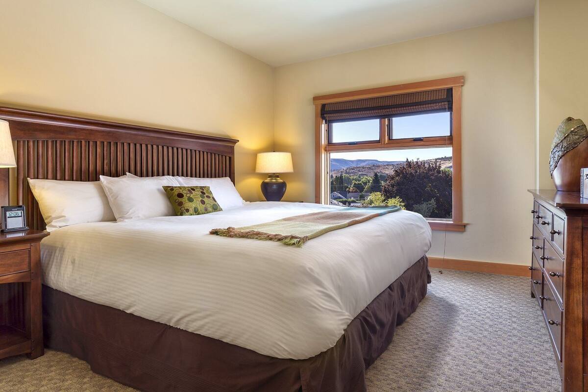 A neatly made bed with a wooden headboard, two bedside lamps, a digital clock, and a dresser with drawers.