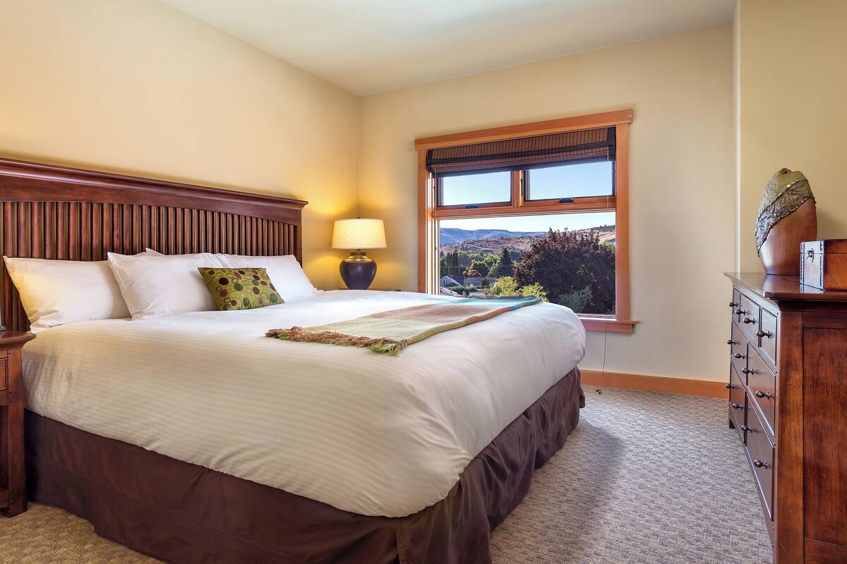 A neatly made bed with white linens and a brown headboard, flanked by two bedside tables with lamps and a digital clock. A window with a view of a mountainous landscape is visible in the background.