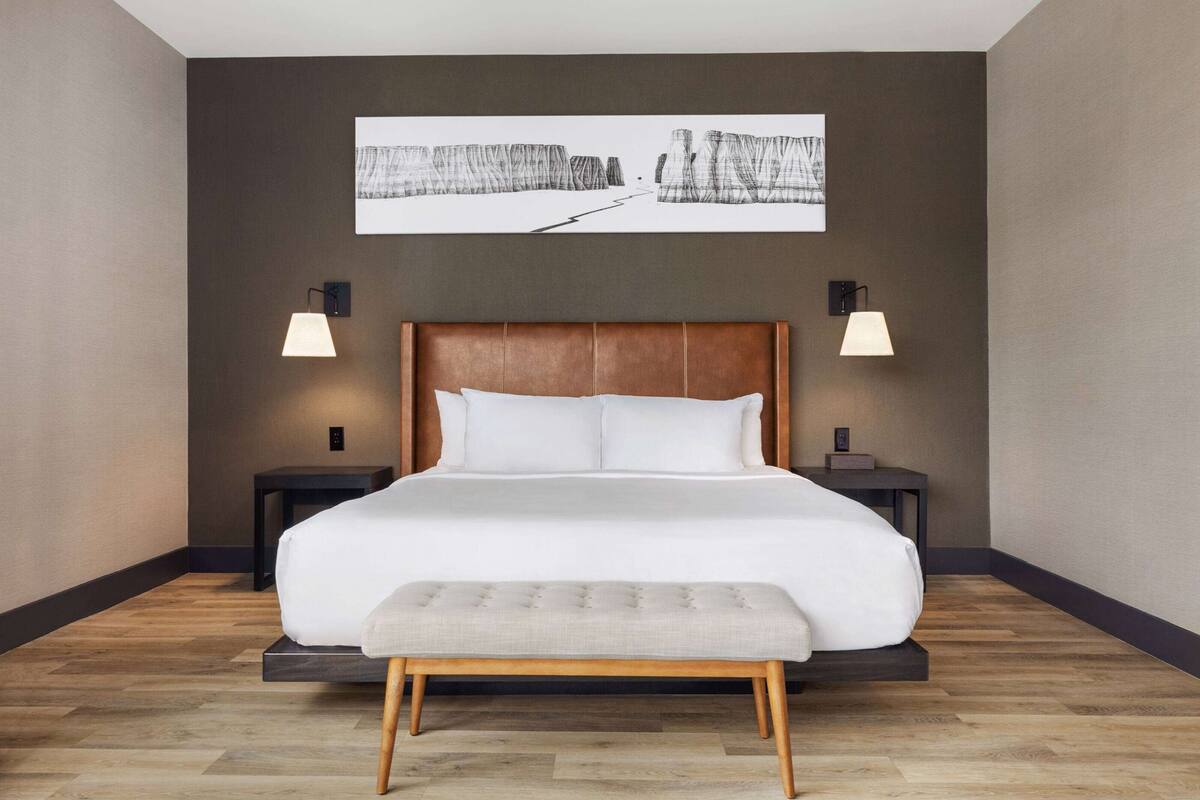 A modern bedroom with a large bed, two bedside tables, a bench, and a wall-mounted artwork.