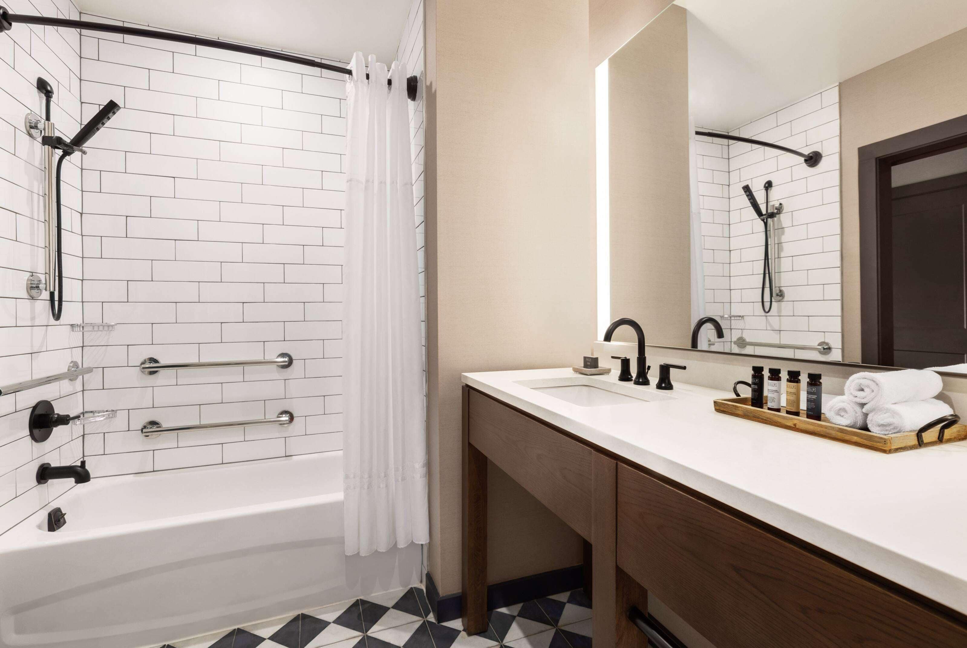 A bathroom with a white-tiled shower, a wooden vanity with a mirror, and a black and white checkered floor.