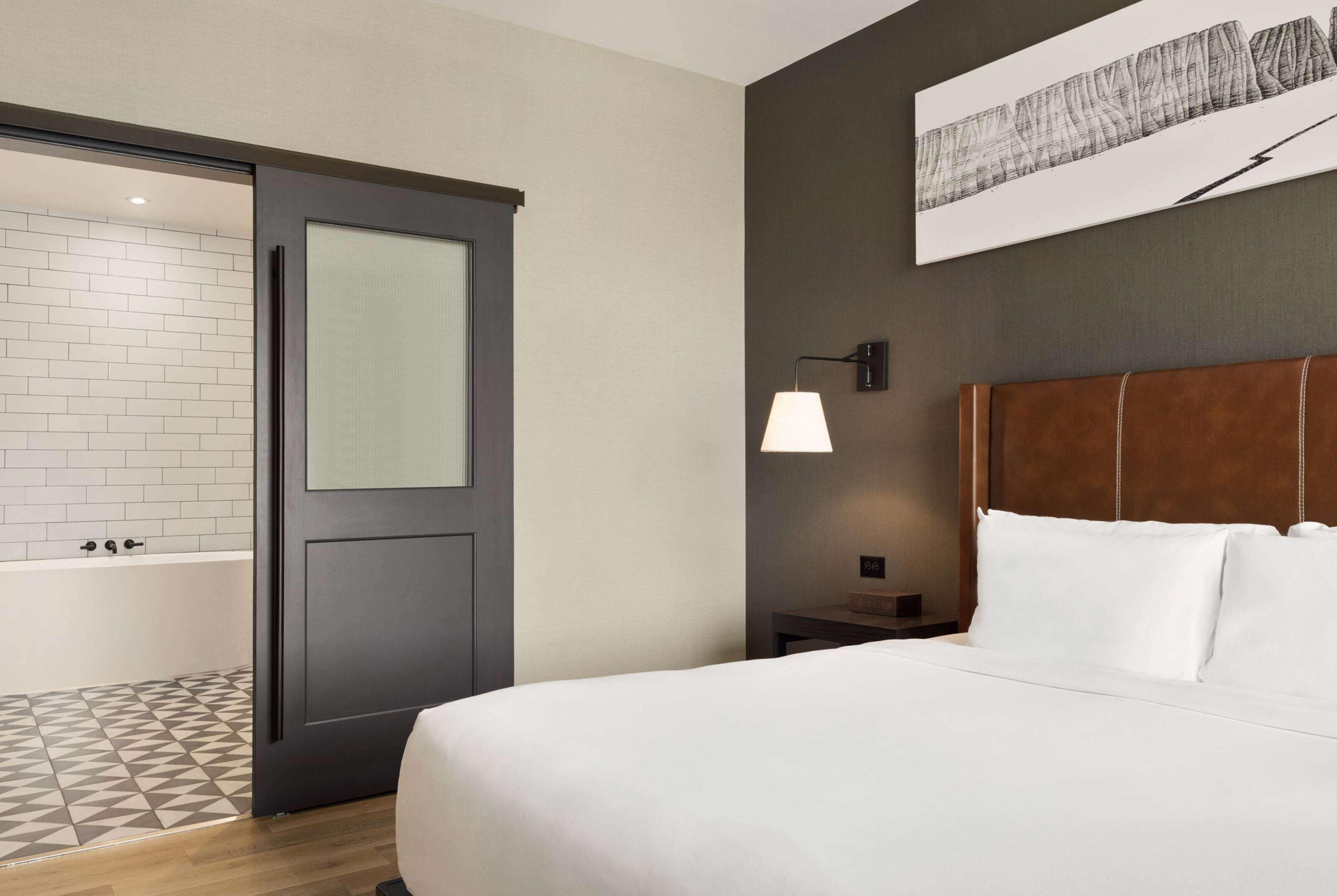 Habitación de hotel con una cama grande, una mesita de noche, una lámpara, una obra de arte en la pared y un baño con bañera y suelo de baldosas.