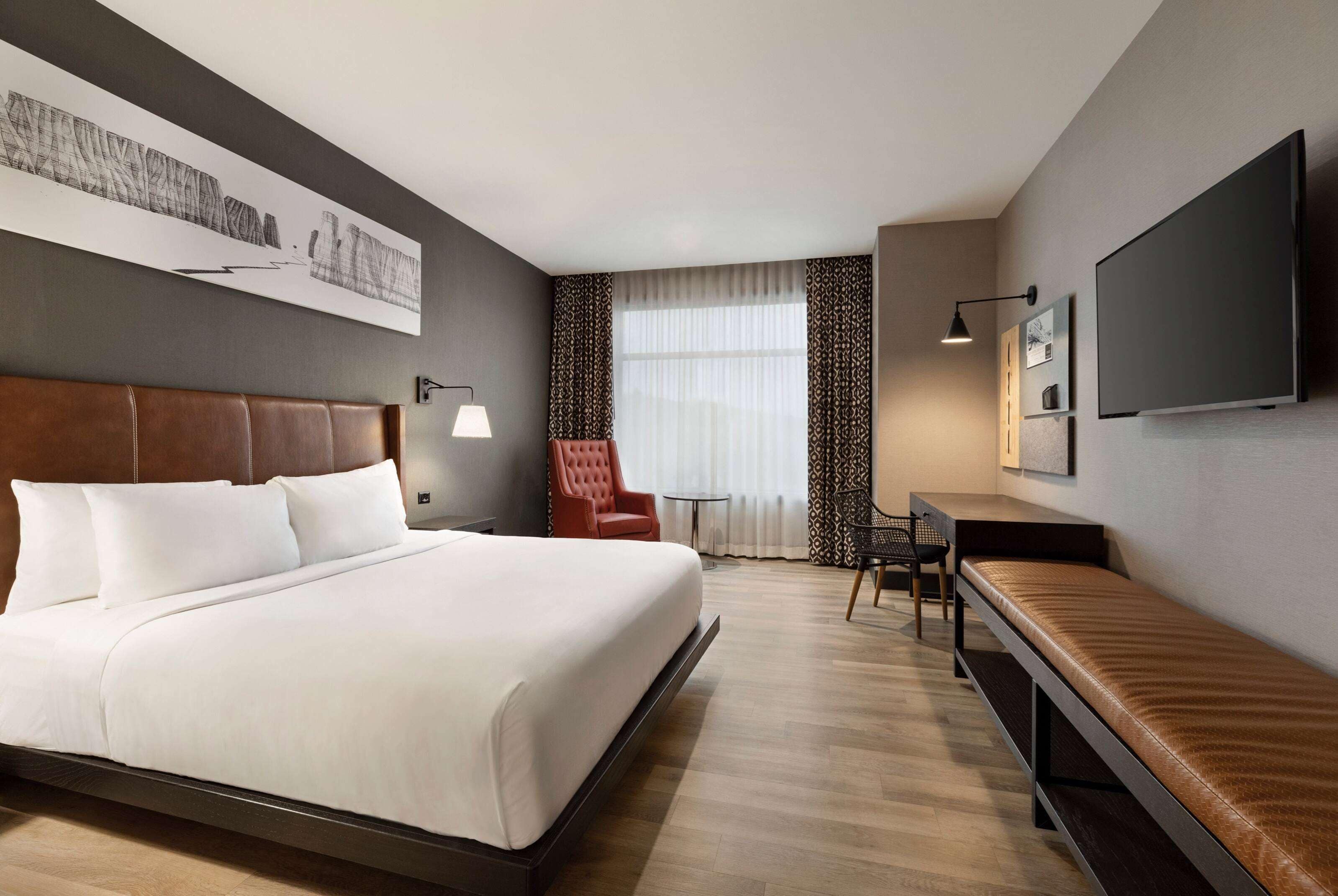 A hotel room with a large bed, a desk, a chair, a TV, and a window with curtains.