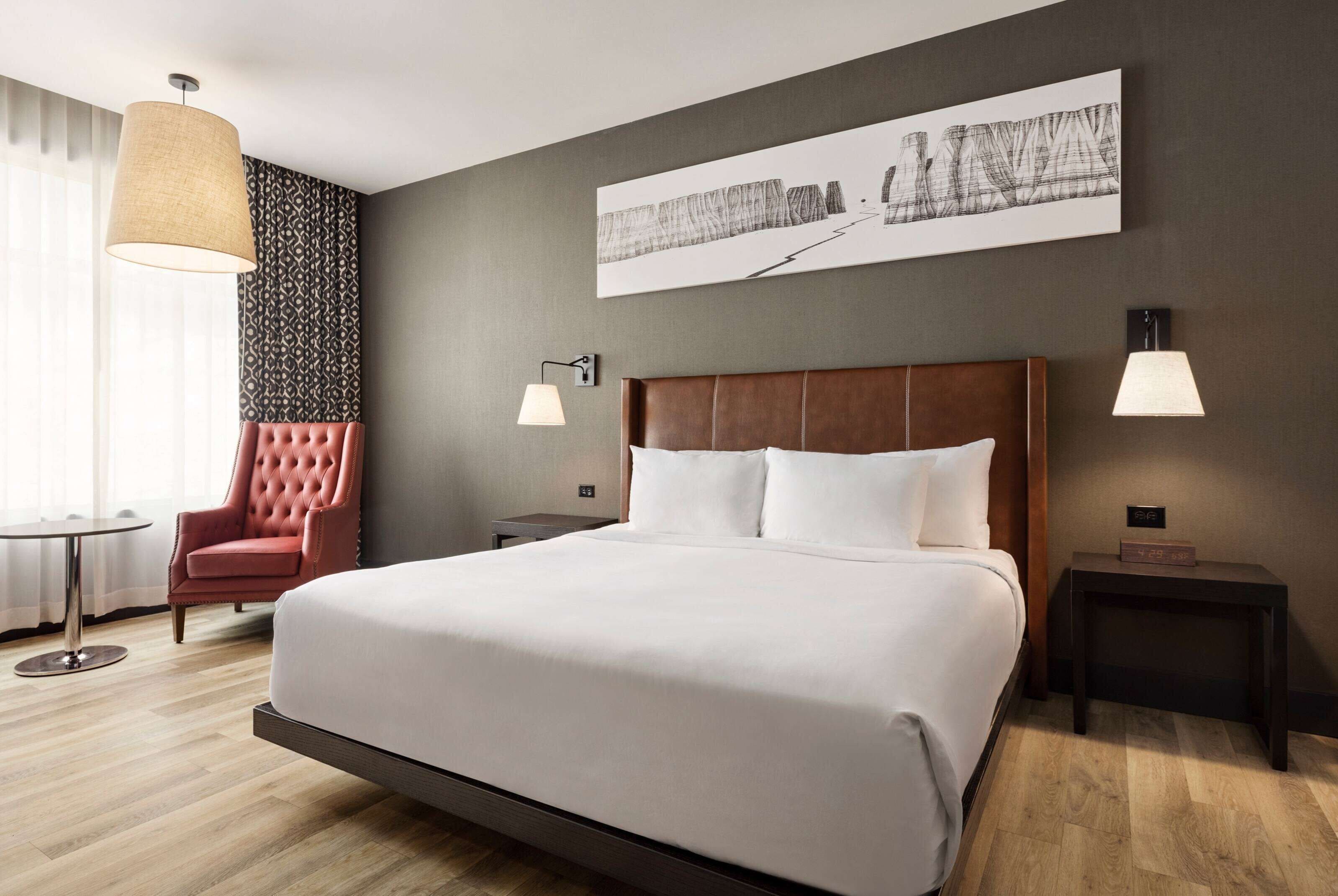 Een hotelkamer met een groot bed, een rode stoel, een klein tafeltje en een kunstwerk aan de muur.