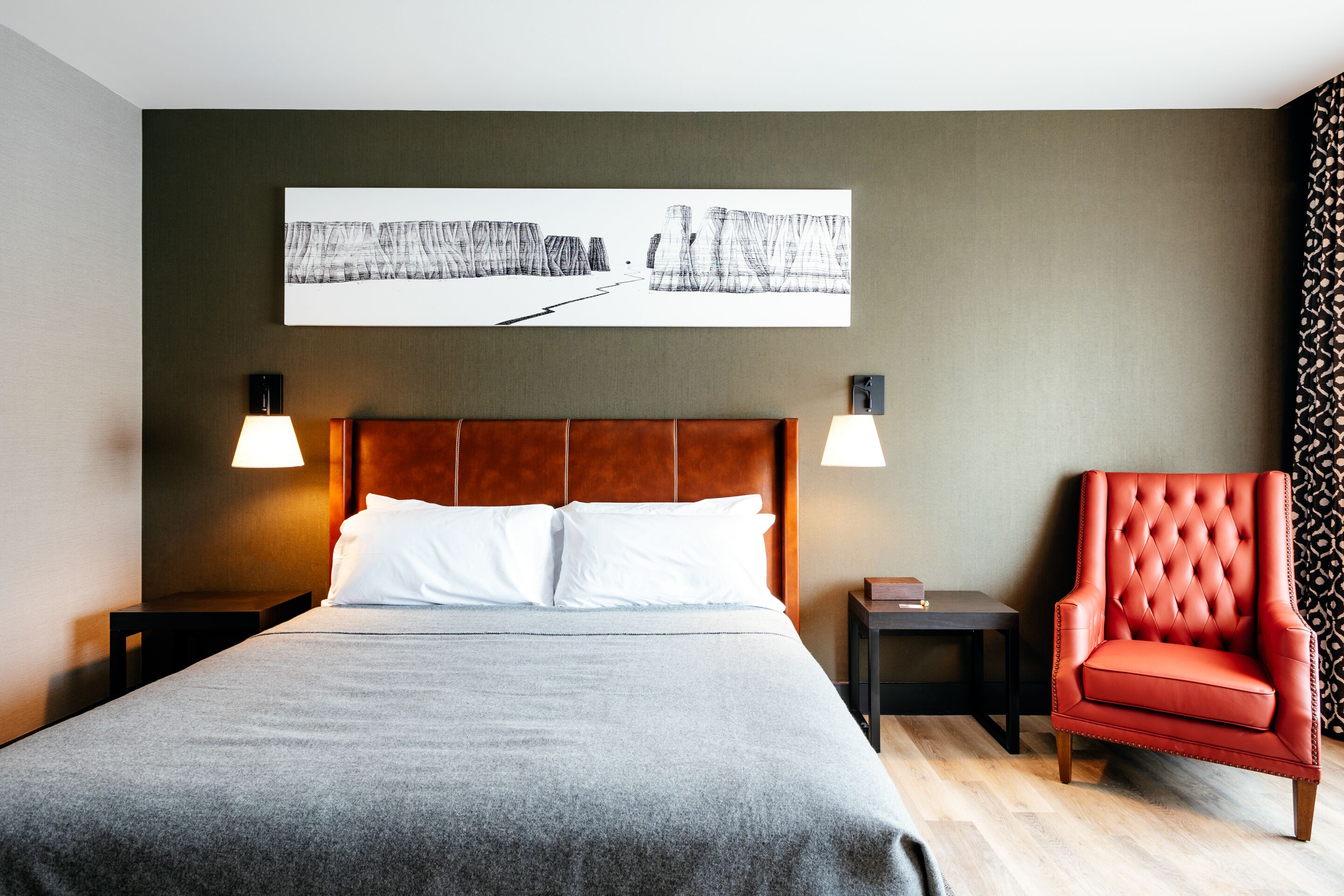 Habitación de hotel con una cama grande, dos mesitas de noche, un sillón rojo y una obra de arte colgada en la pared.