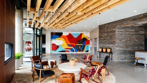 Un vestíbulo moderno con vigas de madera, un mural abstracto y colorido, y una variedad de opciones de asientos, incluyendo sillones de cuero y taburetes de madera.