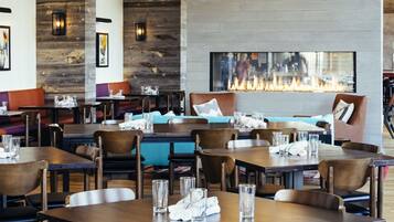 A modern restaurant interior with wooden tables, chairs, and a central fireplace.