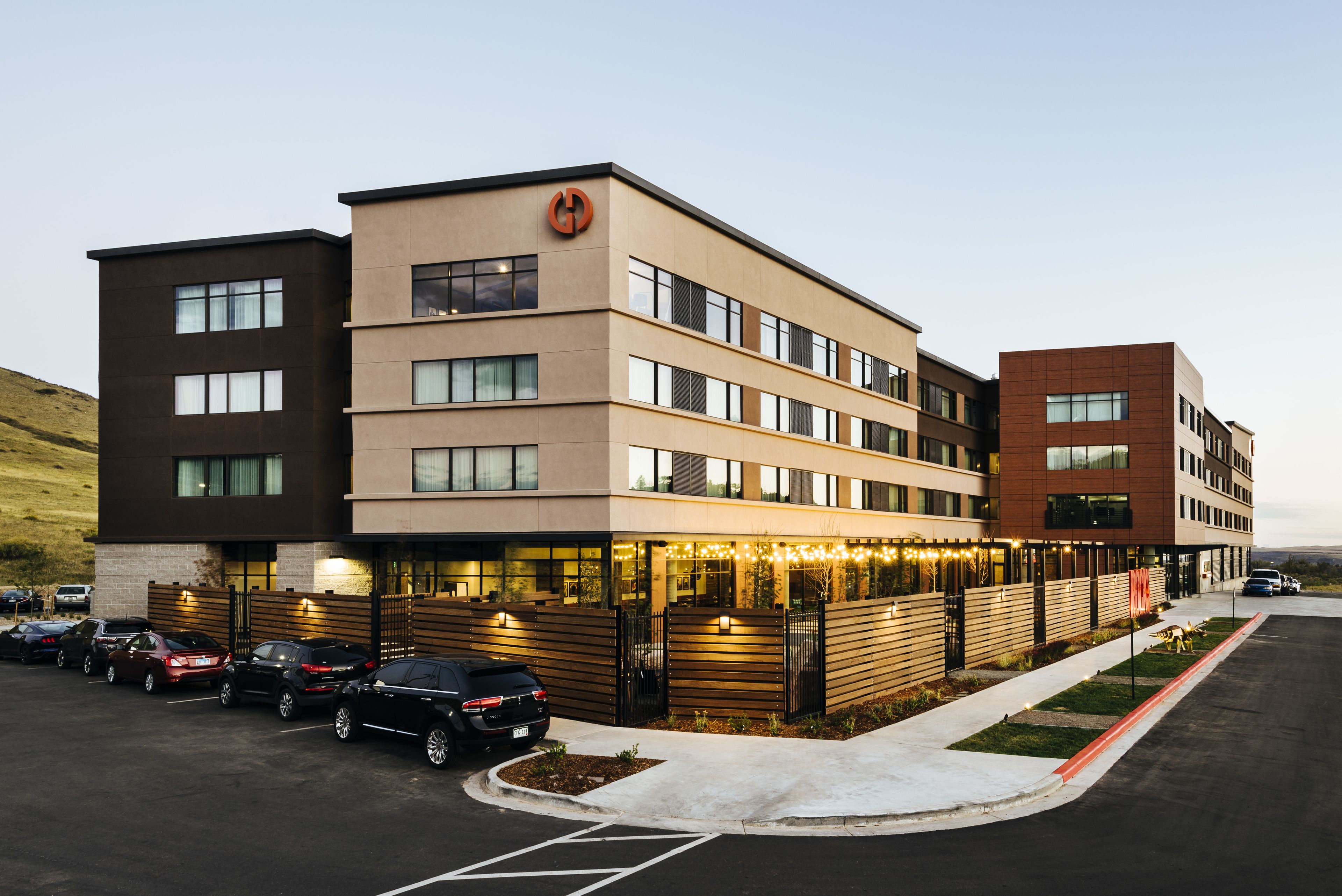 A multi-story hotel with a modern design, featuring a parking lot with several cars and a landscaped area with trees.