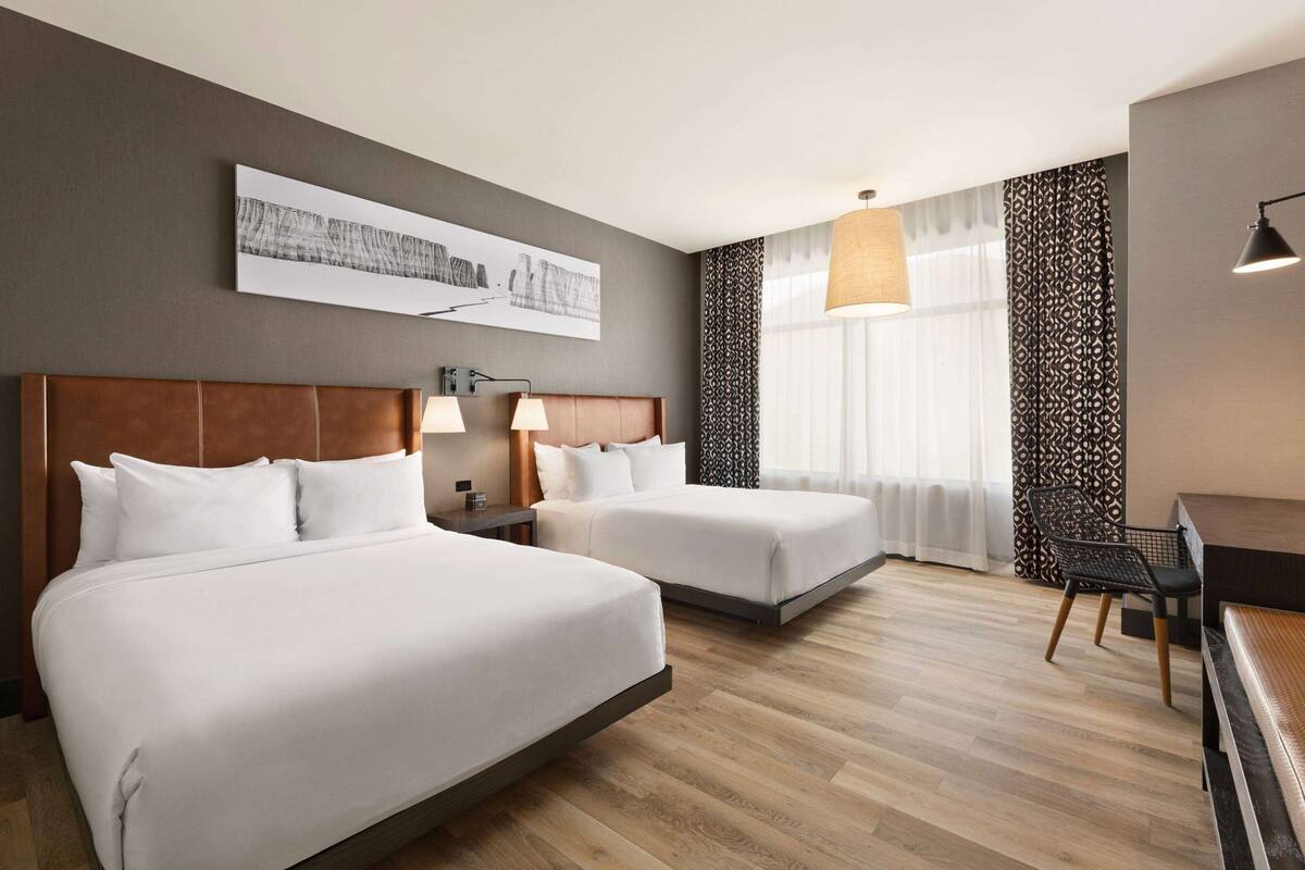 A hotel room with two beds, a desk, a chair, a large window with curtains, and a wall-mounted artwork.