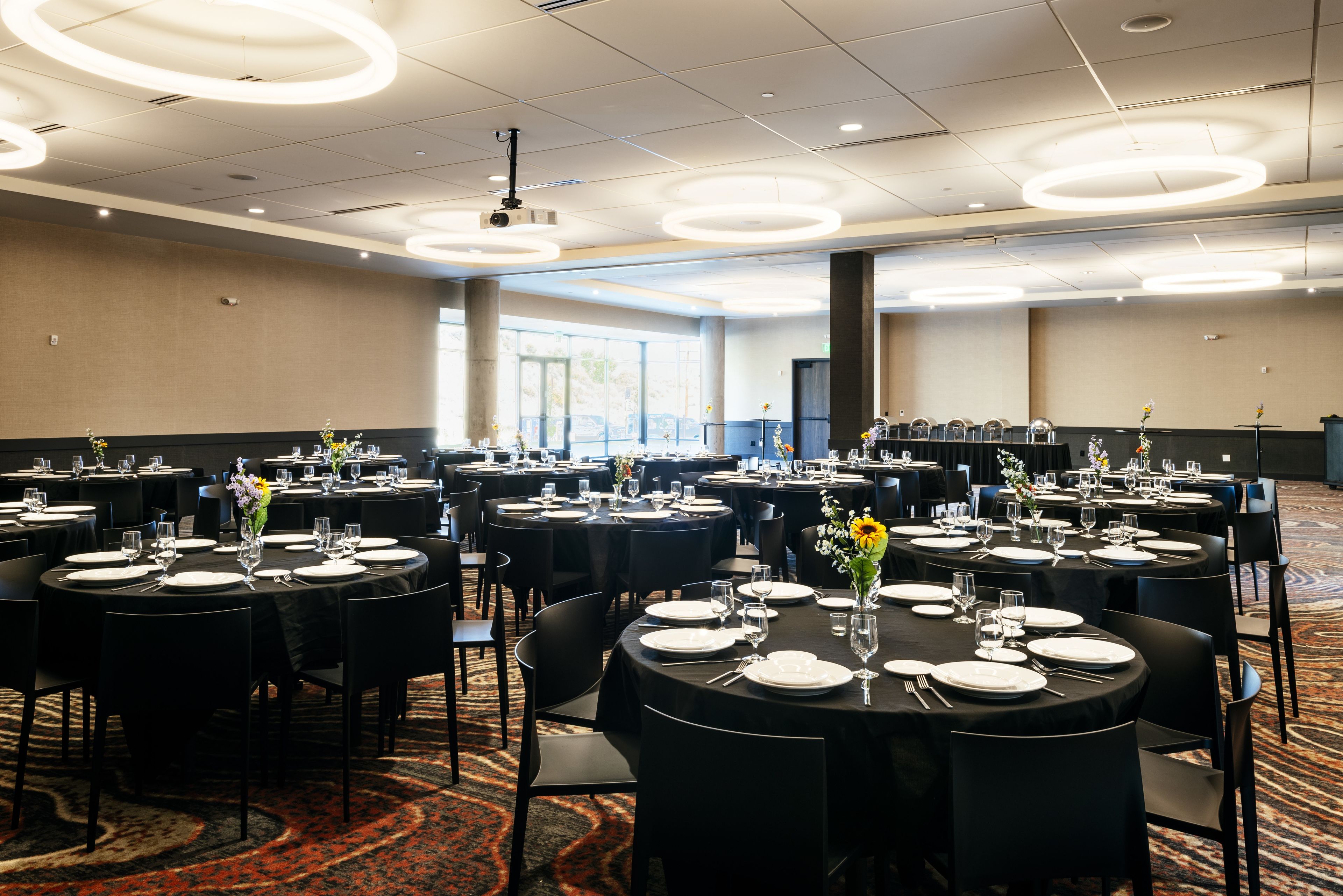 A conference room with multiple round tables, each set with white plates, cutlery, and glassware, and adorned with floral centerpieces.
