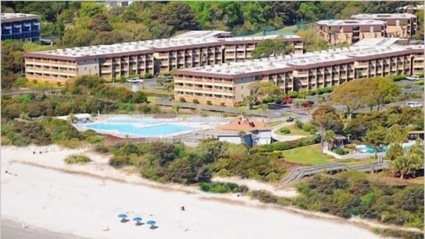 Et stort hotel med mange etager, en swimmingpool og en strand med parasoller.