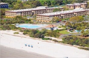 A large hotel with multiple floors, a swimming pool, and a beach area with umbrellas.