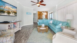A living room with a teal sofa, a white armchair, a wicker coffee table, a ceiling fan, and a large painting of a beach scene. - Breathtaking Views, Views, Views! Oceanfront wraparound balcony; 3 BR and 3 BA. (Orange Beach)