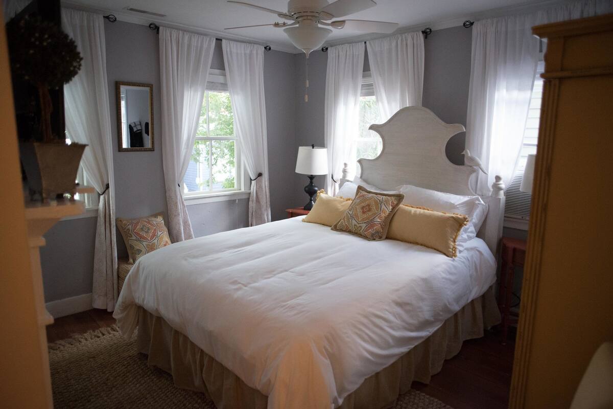 A large bed with white bedding and decorative pillows, a ceiling fan, and a window with sheer curtains.