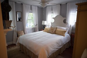 A large bed with white bedding and decorative pillows, a ceiling fan, and a window with sheer curtains.