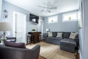 A living room with a grey sofa, a small dining table, a ceiling fan, and a TV mounted on the wall. - Suites on Cannon (Charleston)