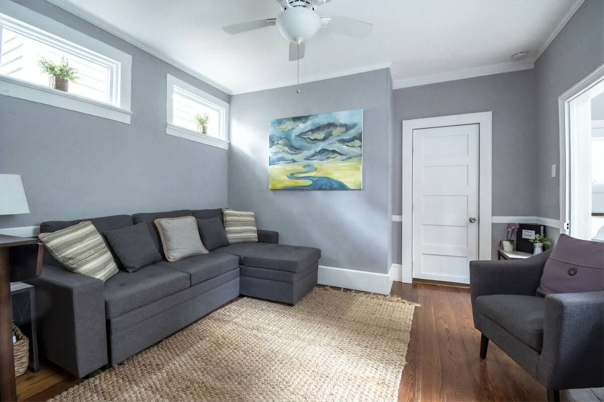 A living room with a grey sectional sofa, a smaller grey armchair, a large painting, and a ceiling fan.