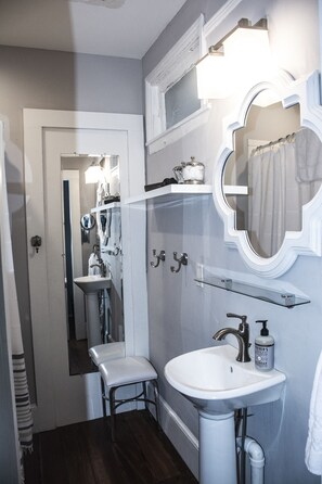 A bathroom with a white sink, a mirror, and a shower curtain. There is a towel rack above the sink and a shelf with toiletries. - Suites on Cannon (Charleston)