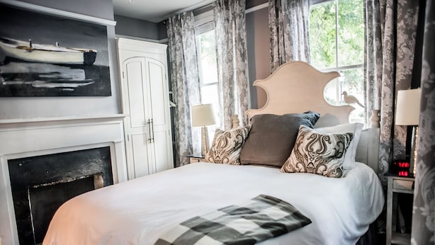 A bedroom with a large bed, a fireplace, a painting, and a window with curtains. - Suites on Cannon (Charleston)