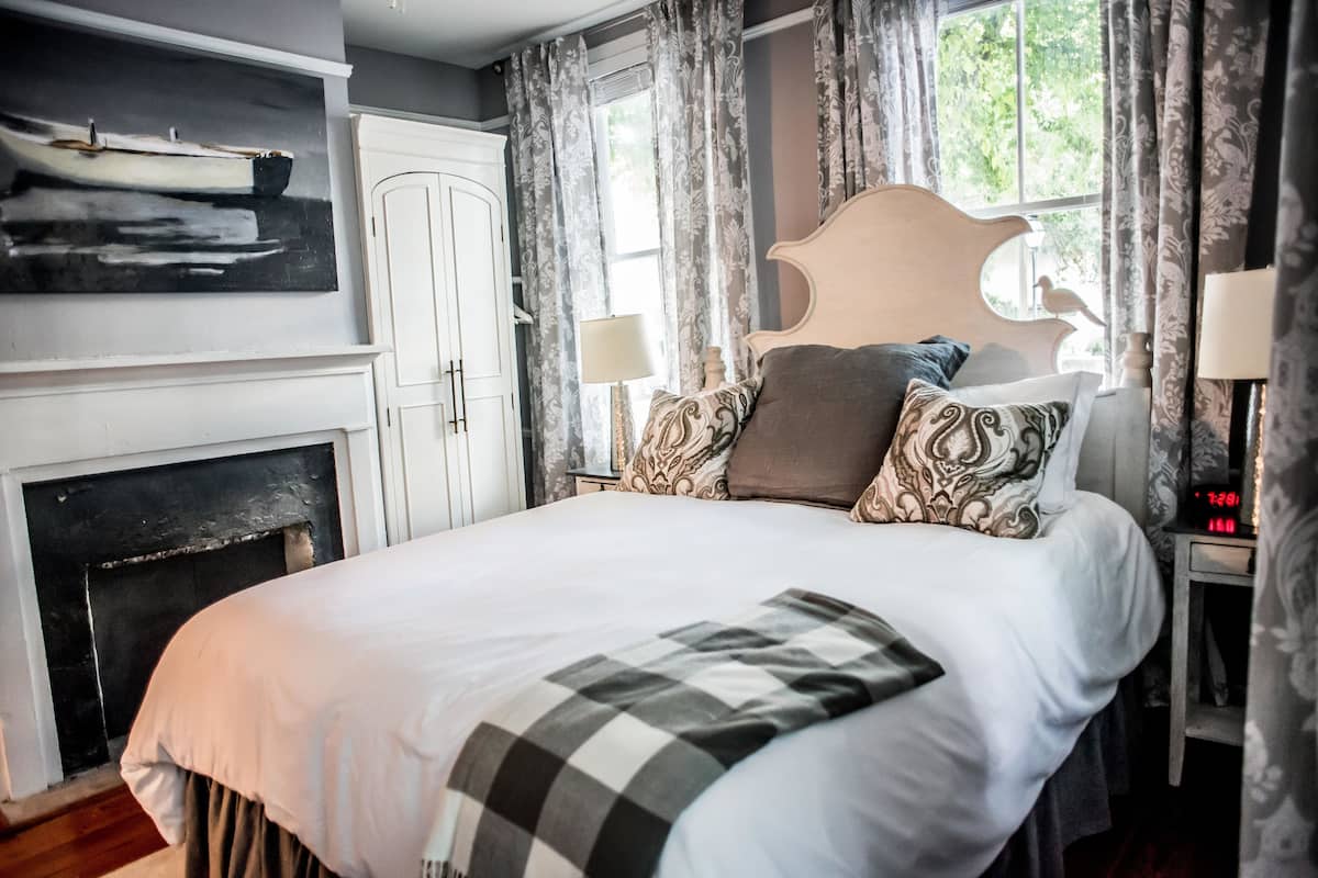 A bedroom with a large bed, a fireplace, a painting, and a window with curtains.