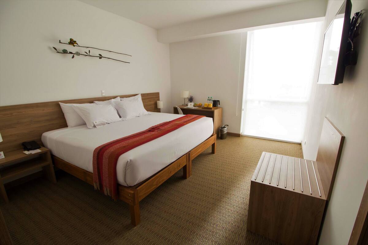Ein Hotelzimmer mit einem großen Bett, einem Nachttisch mit Lampe, einem kleinen Tisch mit Toilettenartikeln und einem an der Wand befestigten Fernseher.