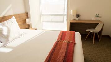 A hotel room with a large bed, a desk, a chair, a lamp, and a window.