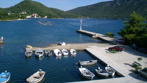 En småbåtshamn med flera mindre båtar förtöjda, en brygga som sträcker sig ut i vattnet och en bakgrund av grönskande kullar.