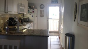 Cocina con gabinetes blancos, microondas, estufa y pileta. Hay un bote de basura y una puerta que da a un balcón.