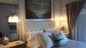 Un dormitorio con cabecera capitonada, una mesita de noche con lámpara, un cuadro sobre la cama y una ventana con cortinas.