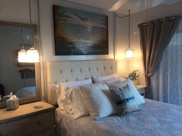 Une chambre à coucher avec une tête de lit capitonnée, une table de chevet avec une lampe, un tableau au-dessus du lit et une fenêtre avec des rideaux.