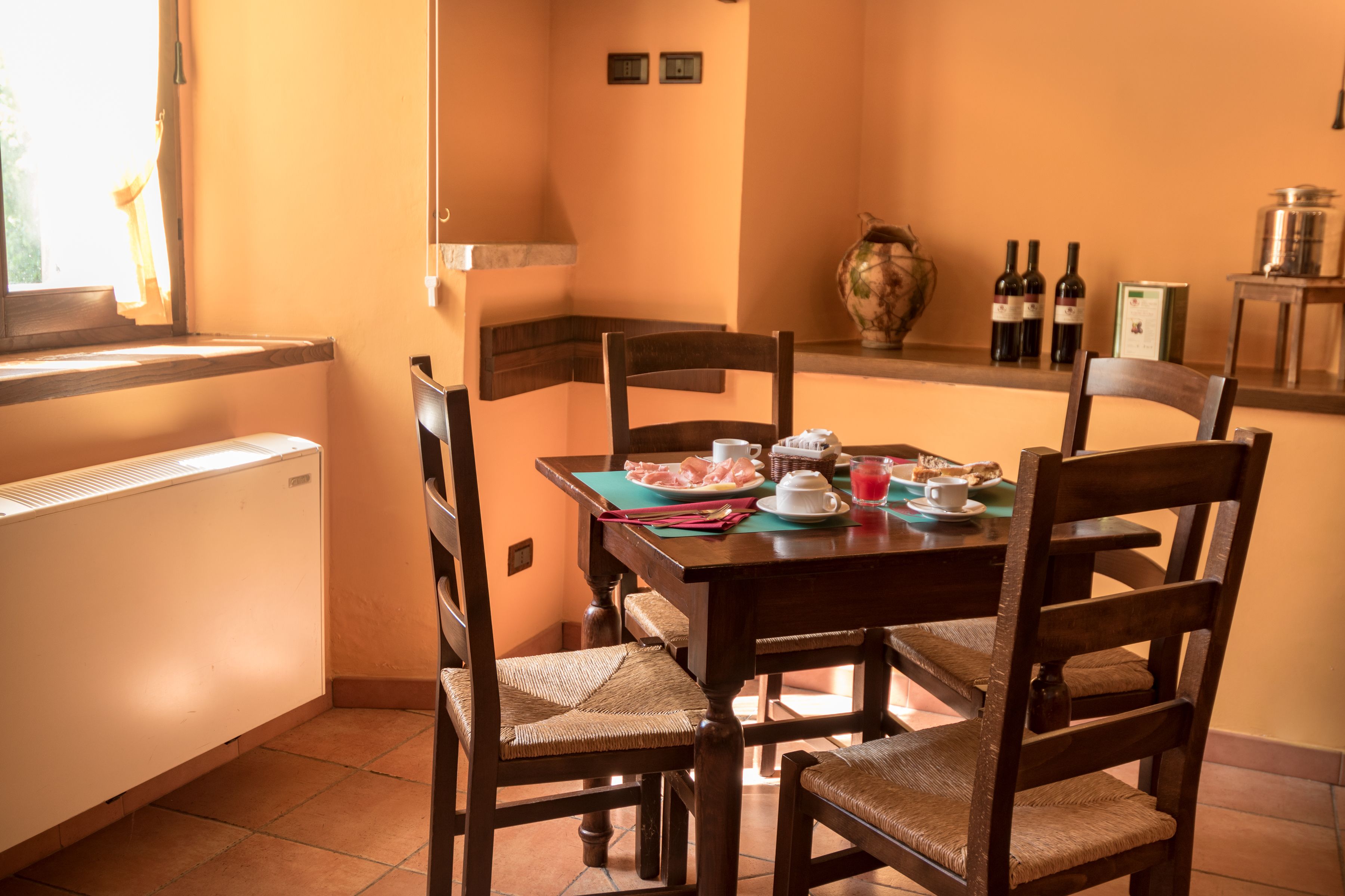 Un comedor con una mesa dispuesta para cuatro personas, rodeada de sillas de madera con asientos de mimbre. En la mesa hay un florero y botellas de vino, y se ve una ventana con cortinas al fondo.