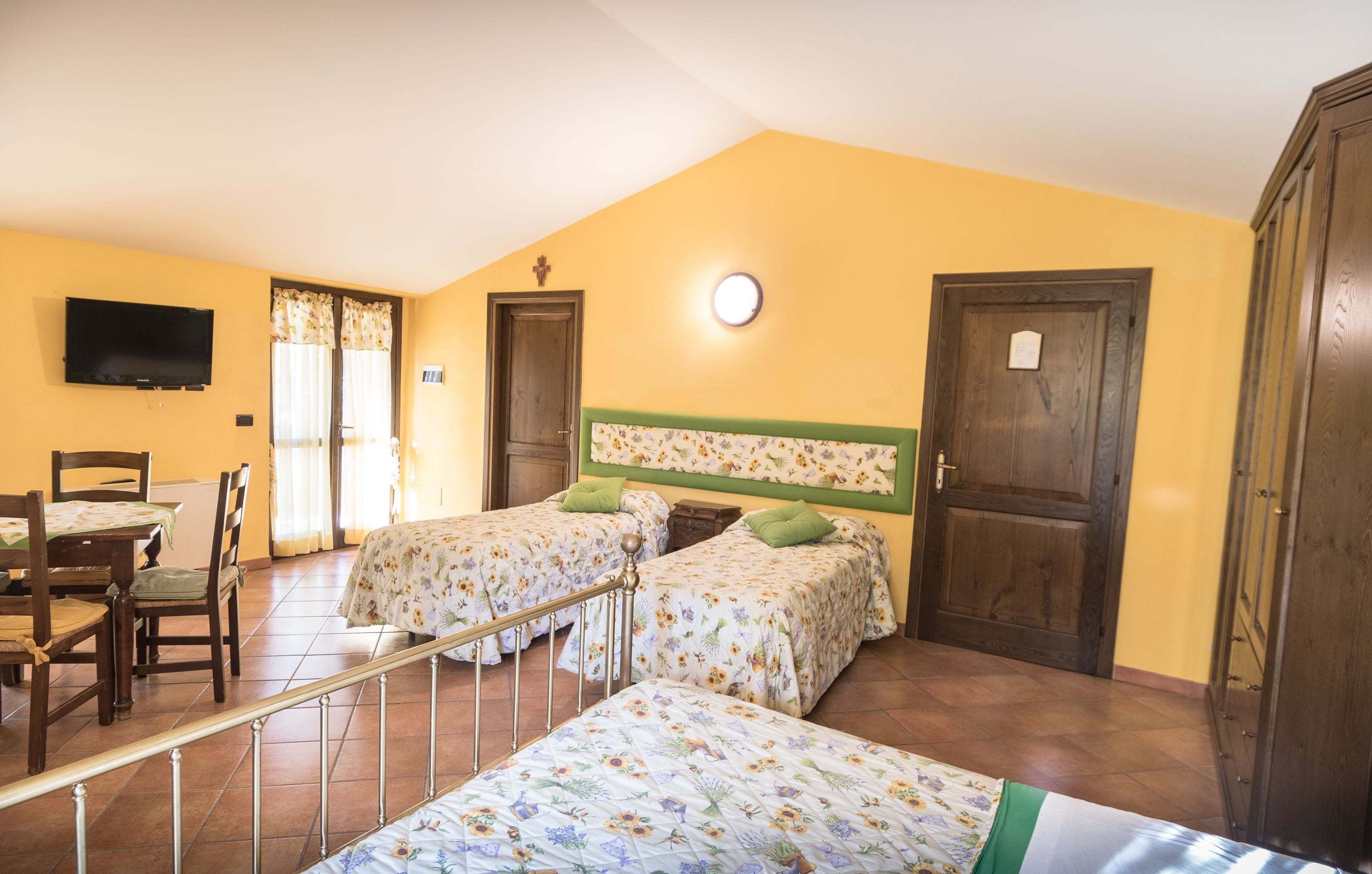 Ein Zimmer mit zwei Betten, einem Esstisch, Stühlen, einem Fernseher und einem Fenster mit Vorhängen.