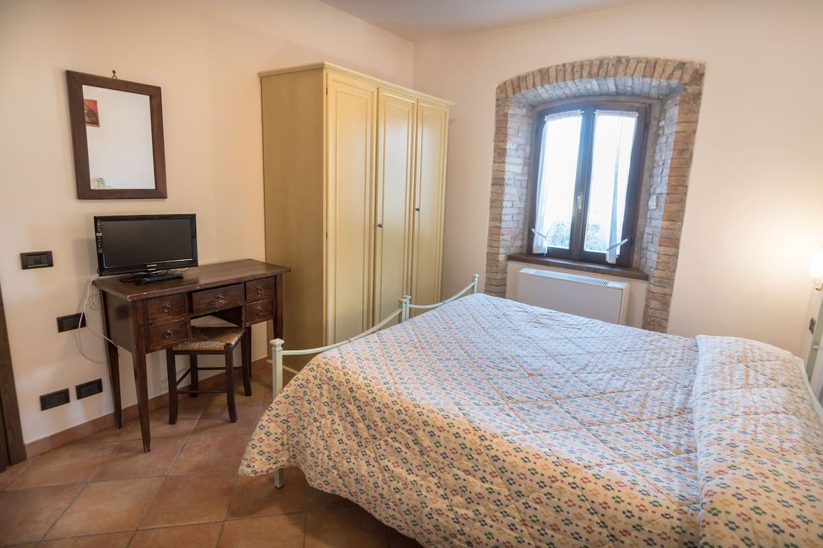 Une chambre à coucher comprenant un lit, un bureau, une chaise, une armoire et une fenêtre surmontée d’une arche en briques.