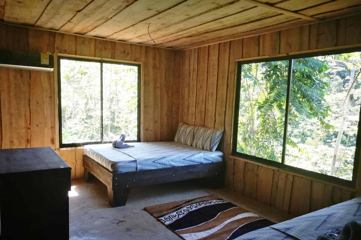 A wooden cabin with a single bed, a small table, and a window with a view of greenery.