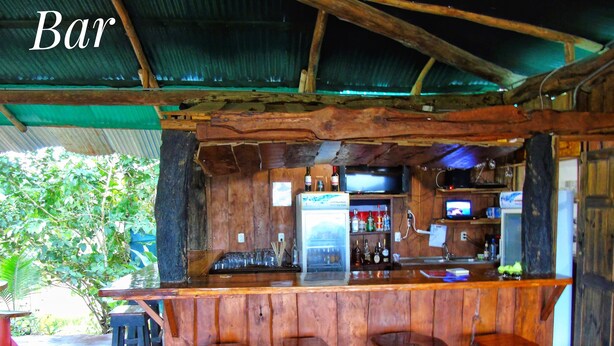 A rustic bar with wooden stools, a refrigerator, and shelves of bottles. - El Tesoro de Sir Hotel (Drake Bay)