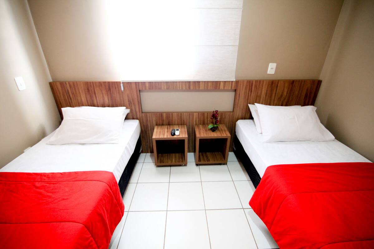 Ein Hotelzimmer mit zwei Einzelbetten, jeweils mit roter Bettdecke, weißen Kissen und einem Holzkopfende. Es gibt zwei Nachttische aus Holz, auf einem davon eine Fernbedienung und eine kleine Pflanze.
