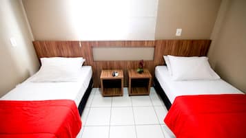 Habitación de hotel con dos camas individuales, cada una con colcha roja, almohadas blancas y cabecero de madera. Hay dos mesitas de noche de madera, una con un control remoto y una planta pequeña.