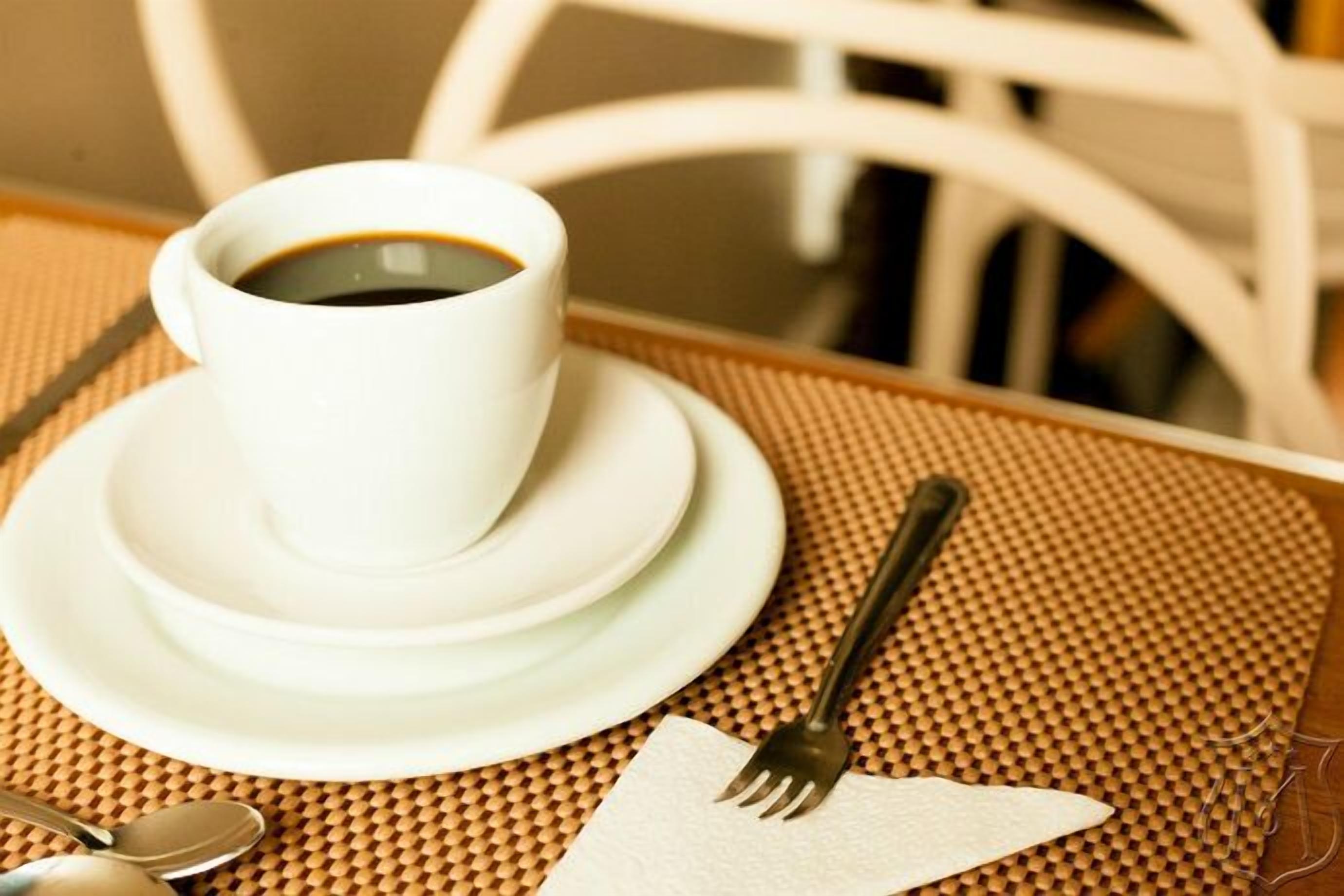 Uma chávena de café branco sobre um pires branco, um garfo e uma colher sobre um apoio de tecido.