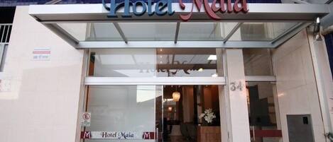 La entrada del Hotel Maia, con fachada de vidrio y una recepción visible en el interior.
