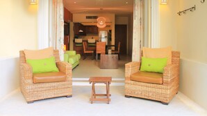 A room with two wicker chairs with green cushions, a small wooden table, and a view into a dining area with a ceiling fan. - Luna Maya Beautiful 2BR Beach Condo (Playa del Carmen)
