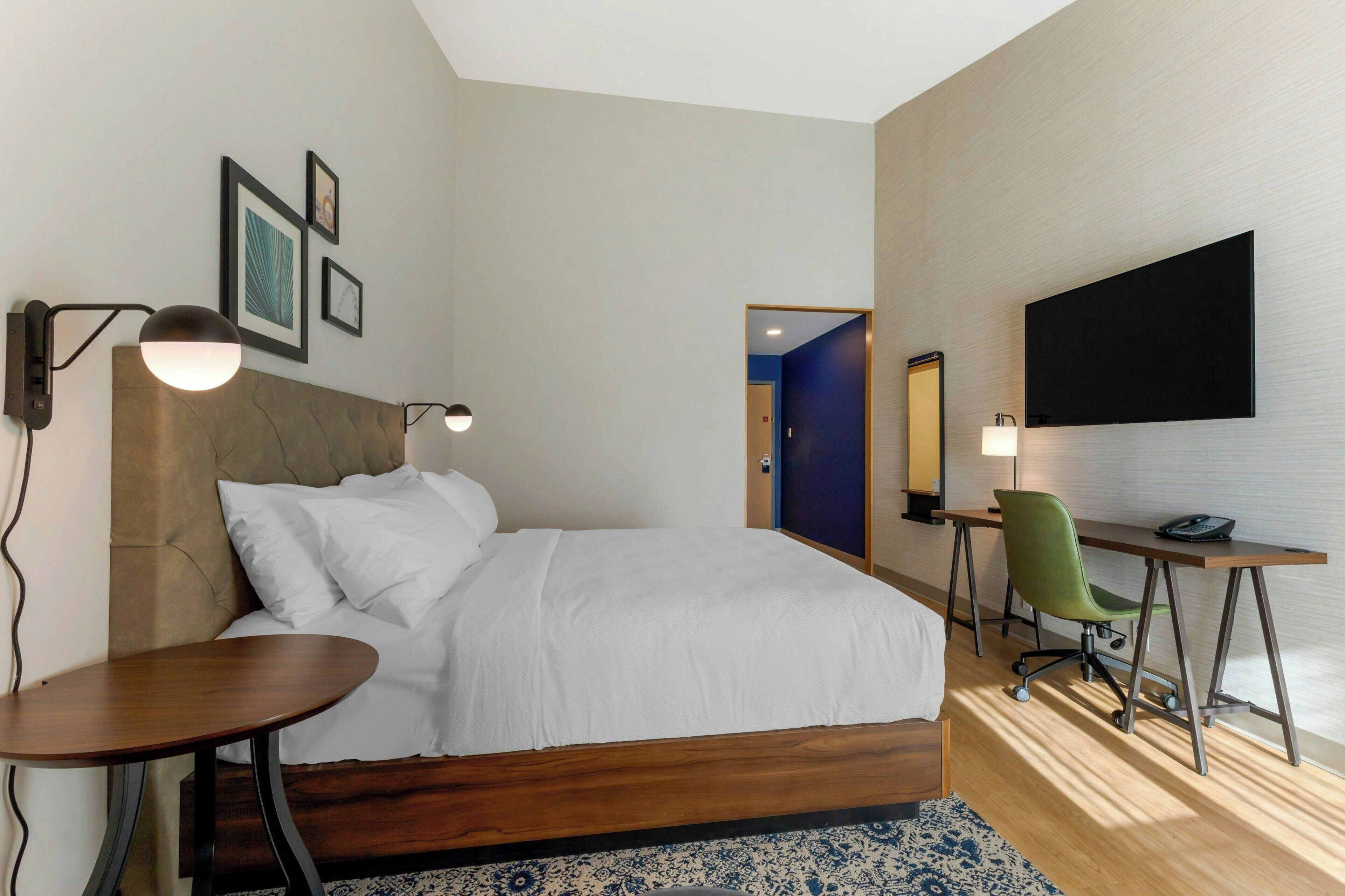 A modern hotel room with a large bed, a desk, a chair, and a TV mounted on the wall.