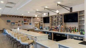 Uma área de bar com um balcão comprido, vários bancos altos, uma televisão na parede e diversas garrafas e copos expostos em prateleiras.