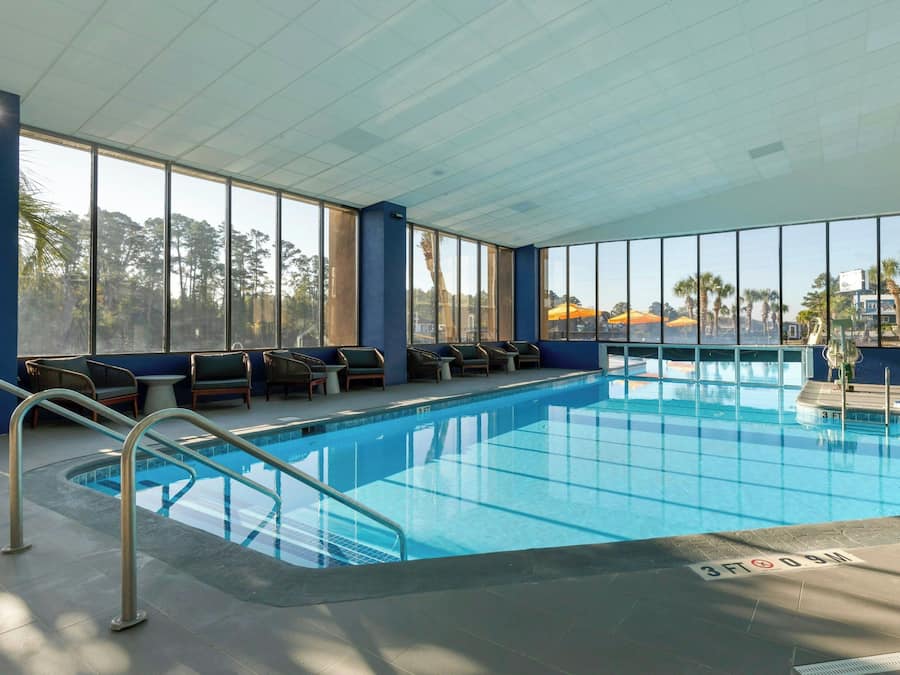 An indoor swimming pool with clear blue water, surrounded by lounge chairs and a view of palm trees through large windows.