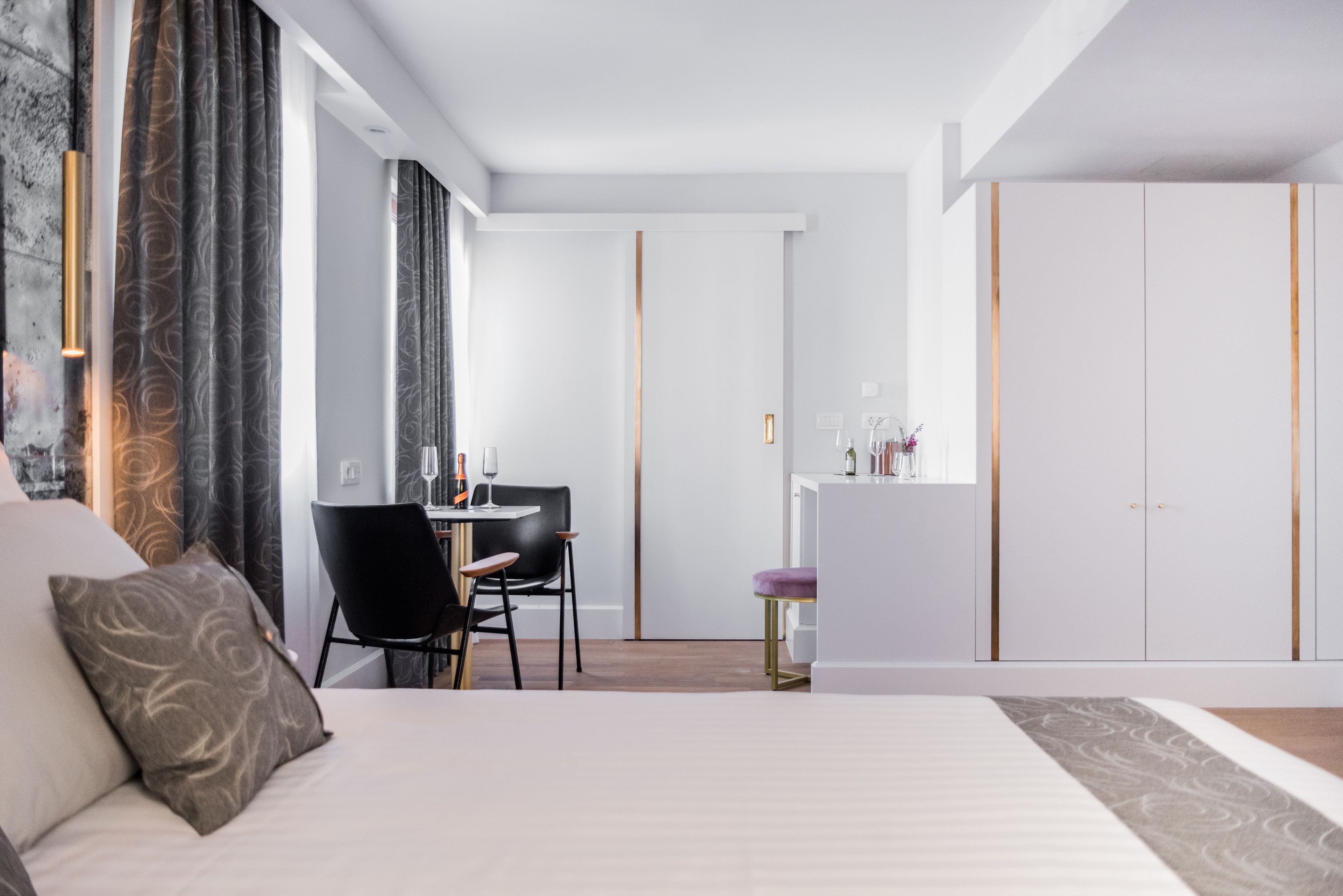 A modern hotel room with a large bed, a desk with a chair, a small stool, and a wardrobe.
