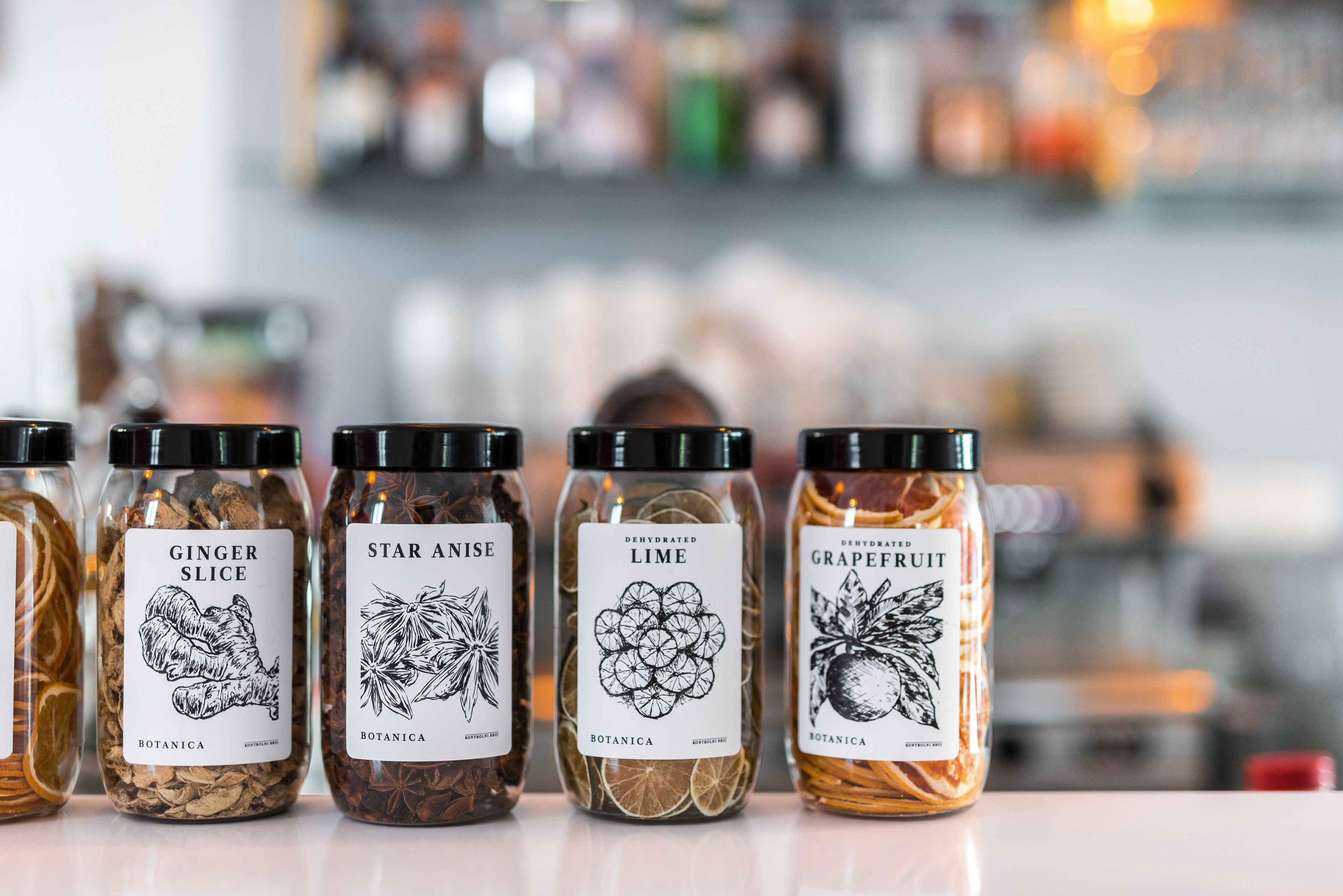 Quatre bocaux en verre, étiquetés respectivement « tranches de gingembre », « anis étoilé », « citron vert déshydraté » et « pamplemousse », sont posés sur un comptoir.