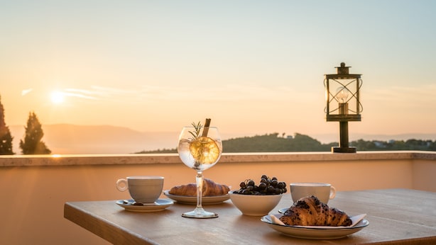A table with a croissant, a bowl of grapes, a glass of wine, and a cup, set against a sunset view with a lantern in the background. - La Loggia Diffused Hotel (Labin)