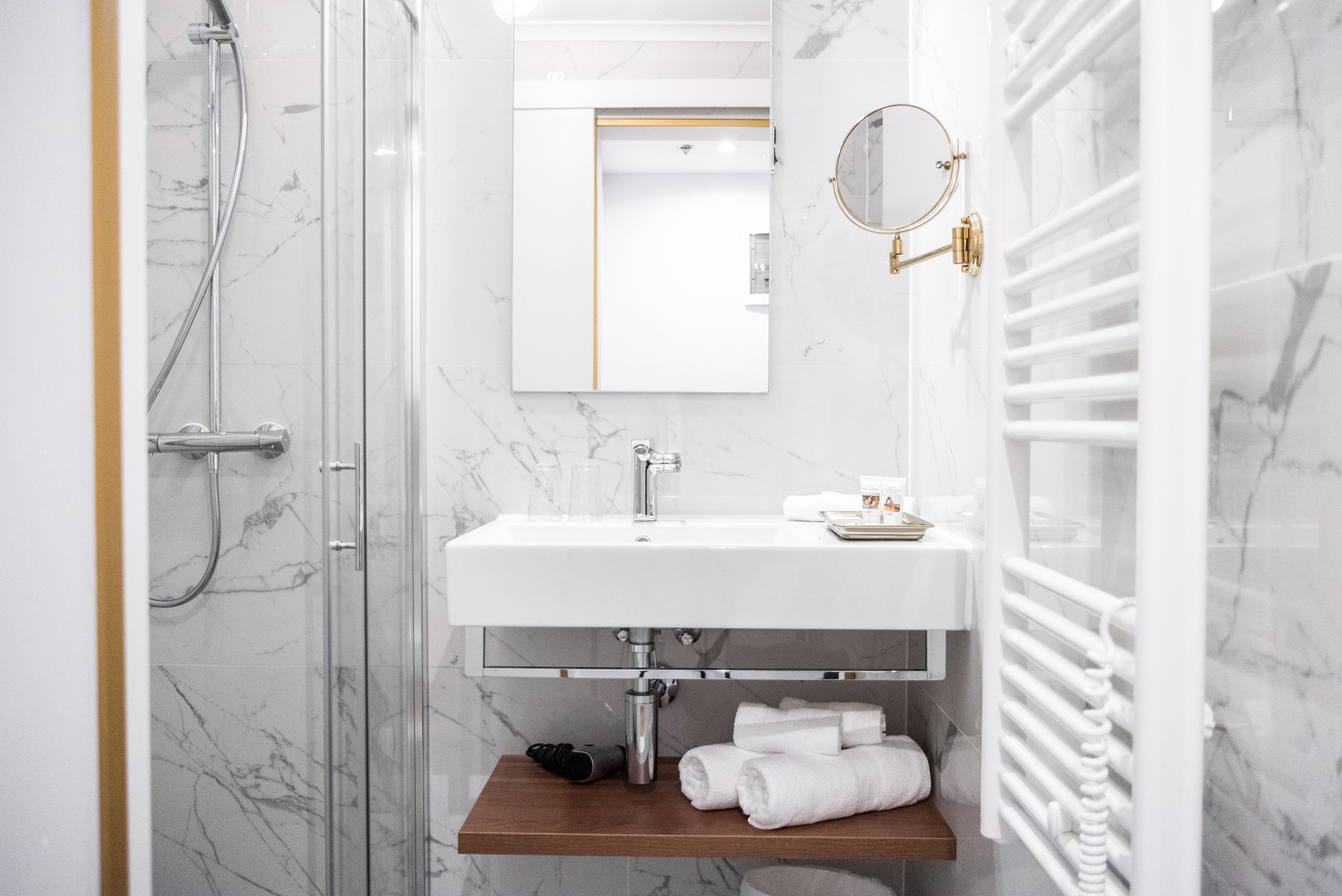Un baño moderno con cabina de ducha de vidrio, un lavabo blanco con grifo dorado y un estante de madera con toallas.